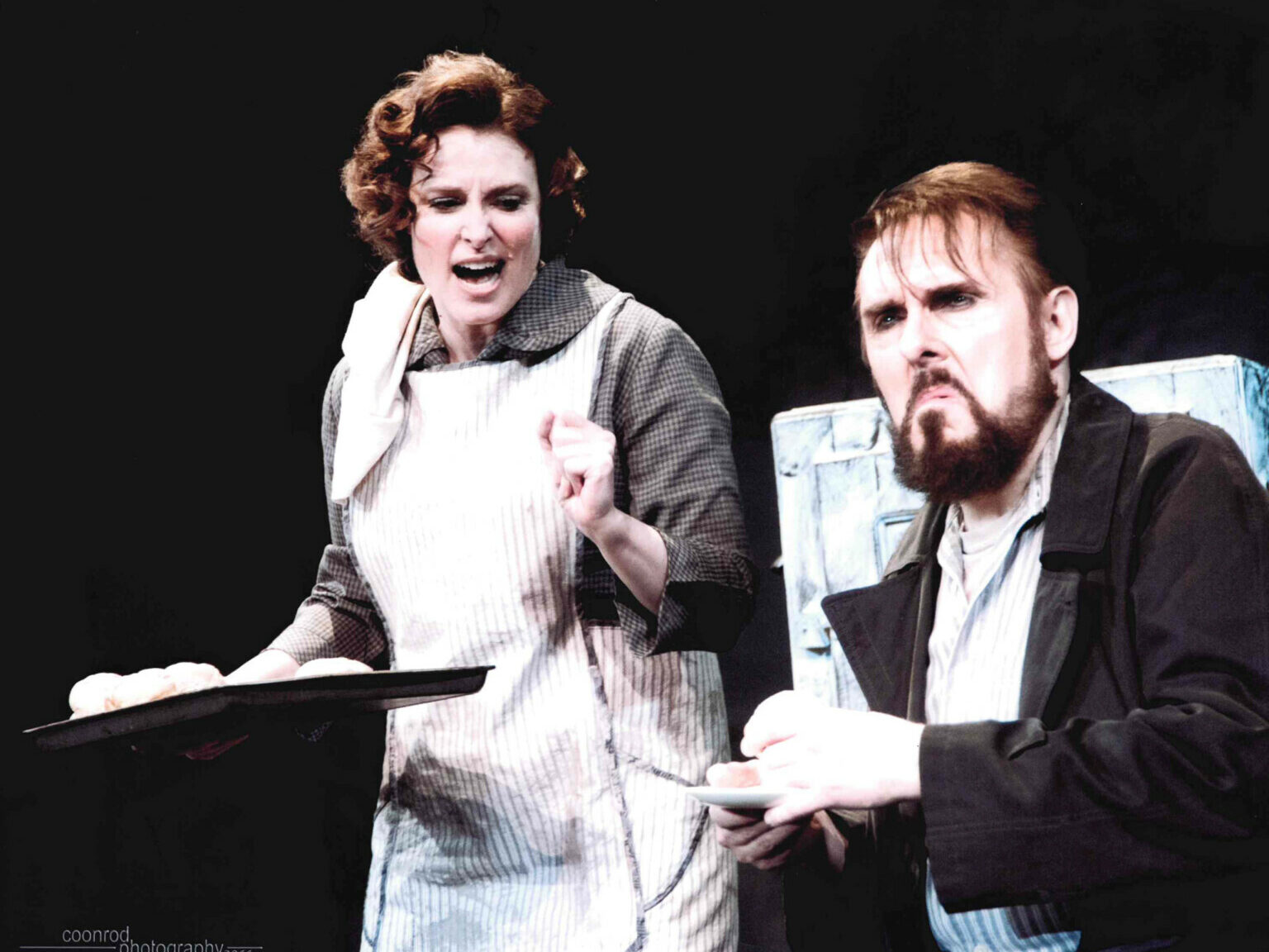 Two actors performing in Sweeny Todd. A woman stands holding a tray of baked goods, while a man sits with a cup of tea in his hands.